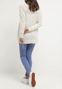 Jasnoszary, teksturowany sweter z długim rękawem, zestawiony z dopasowanymi niebieskimi dżinsami i białymi trampkami. Widok od tyłu.
