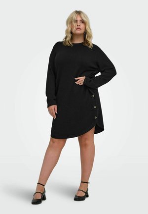 Femme aux cheveux blonds portant une longue robe-pull noire avec des boutons sur le côté et des talons noirs à bride à la cheville, debout devant un fond uni.