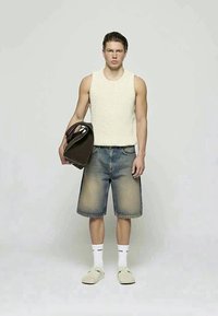 Ung man iklädd en ärmlös krämfärgad stickad väst, långa denimshorts, vita strumpor och tofflor, hållande en stor mörkbrun väska vid sin sida.