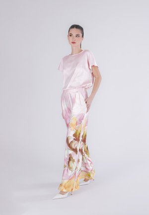 Donna che indossa una maglietta rosa di seta a maniche corte e pantaloni larghi a stampa floreale con tacchi alti bianchi, in piedi davanti a uno sfondo chiaro e semplice.