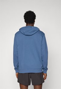 Sudadera con capucha azul y mangas largas, hecha de un material suave, con ribetes en el dobladillo y los puños, combinada con pantalones cortos oscuros.