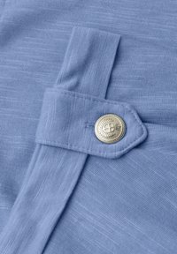 Gros plan sur un tissu bleu clair avec une sangle cousue fixée par un bouton rond argenté orné d'un emblème décoratif.