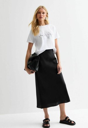 T-shirt graphique blanc avec l'imprimé "FRANCISCO", associé à une jupe midi en satin noire, un sac en cuir noir et des sandales noires.