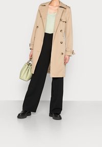 Trench coat beige con bottoni dorati, ampio colletto; abbinato a una maglietta verde chiaro e pantaloni neri a gamba larga. Accessoriato con una piccola borsa verde.