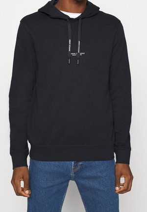 Schwarzer Hoodie mit weißem Armani Exchange-Logo auf der Brust, über einem weißen Hemd getragen, kombiniert mit blauen Jeans bei einer Person.