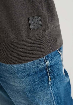 Suéter de punto gris oscuro con dobladillo acanalado y un parche de logo negro; combinado con jeans de mezclilla azul claro con costuras en contraste.