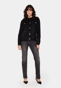 Svart glittrig kofta med guldknappar, strukturerad stickning, rund hals, bärs över grå denimjeans, parat med svarta spetsiga klackar.