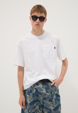 Jonge man met zwarte zonnebril, een wit T-shirt met borstzakje en blauwe en grijze camouflagebroek met één hand in de zak.
