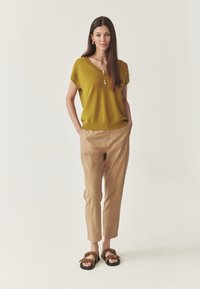 Femme se tenant debout avec les mains dans les poches, portant un pull à manches courtes jaune moutarde, un pantalon beige, des sandales marron et des bijoux en or, sur un fond uni.