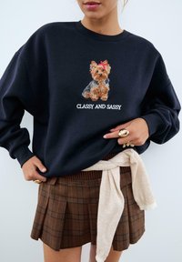 Felpa blu marina con un Yorkshire Terrier ricamato e un fiocco rosso, sopra il testo "CLASSY AND SASSY." Abbinata a una gonna a quadri marrone.