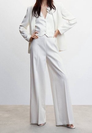Žena oblečená v bílém na míru ušitý oblek se širokými kalhotami, odpovídající vestou, sako přehozené přes ramena a sandály s otevřenou špičkou.