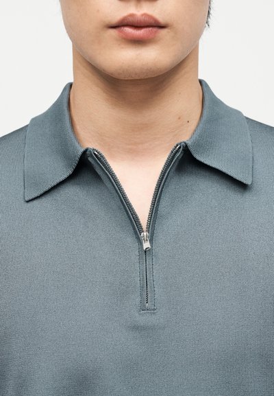 Κοντινό πλάνο ενός ατόμου που φοράει γκρι πλεκτό πόλο πουκάμισο με μερικώς φερμουάρ ασημί στο γιακά.