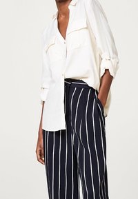 Chemise blanche à boutons avec deux poches poitrine et manches retroussées, associée à un pantalon large rayé bleu marine.