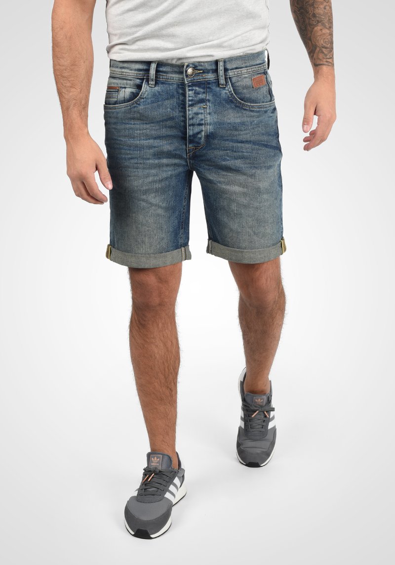 Shorts en denim dans un délavage bleu clair avec des ourlets roulés, dotés d'une fermeture à bouton et fermeture éclair, ainsi que des poches avant et arrière. Associés à des baskets grises.