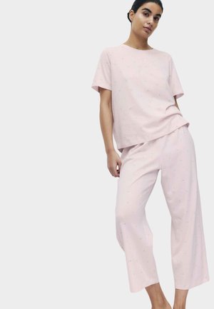 Kobieta ubrana w jasnoróżową bluzkę z krótkim rękawem i pasujące szerokie spodnie z drobnymi, subtelnymi wzorami kwiatowymi, stojąca na białym tle.