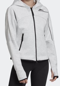Sweat à capuche zippé Adidas gris clair avec fermeture éclair noire, poches zippées, trous pour les pouces et leggings noirs portés par une personne du cou aux hanches.