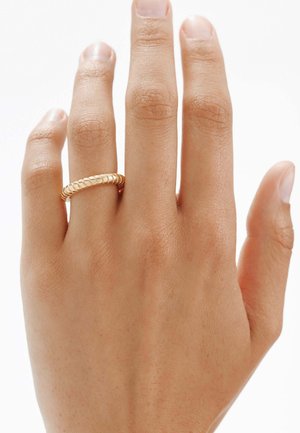 Anillo de oro en una mano, con un diseño suave y redondeado y acentos estriados. El tono de piel es medio con un acabado natural.