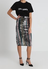T-shirt noir avec le texte "CRY HARD" ; jupe crayon à sequins multicolores avec des rayures verticales ; chaussures à talons noirs avec des brides autour de la cheville.