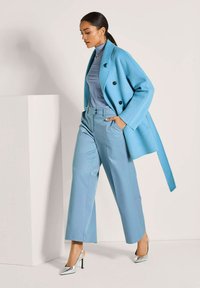 Vrouw die loopt in lichtblauwe wijde broek, bijpassende dubbel-breasted jas, coltrui, zilveren hakken, grote hoepeloorzouders en kettingen.