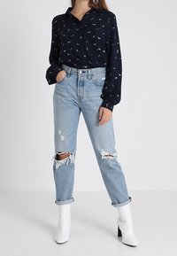 Donna che indossa una blusa a fantasia blu navy, jeans strappati di un leggero blu con risvolti e stivaletti bianchi con tacco, in piedi con una mano nella tasca.