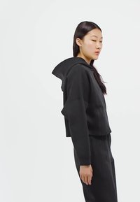 Woman with long dark hair wearing a black cropped hoodie and black pants, facing sideways against a plain white background.