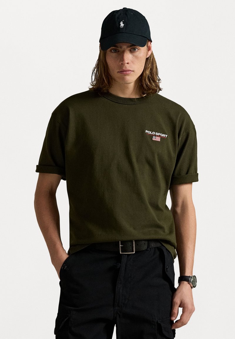 Olivgrünes Baumwoll-T-Shirt mit umgeschlagenen Ärmeln und "POLO SPORT"-Logo. Schwarze Mütze mit gesticktem Logo. Schwarze Hose mit Gürtel und Uhr.
