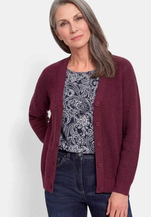Donna matura con capelli grigi indossa una maglietta bianca e nera con motivo, un cardigan bordeaux con bottoni e jeans blu scuro, in piedi contro uno sfondo uniforme.