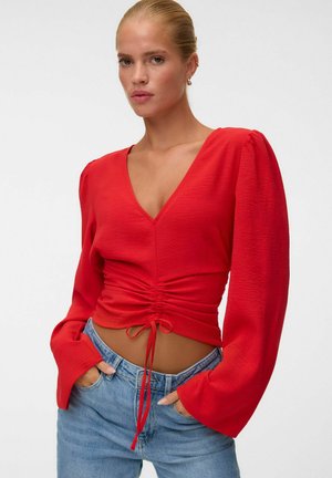 Femme aux cheveux blonds portant un haut rouge à col en V froncé à manches longues et un jean bleu, les mains dans les poches, sur un fond blanc uni.