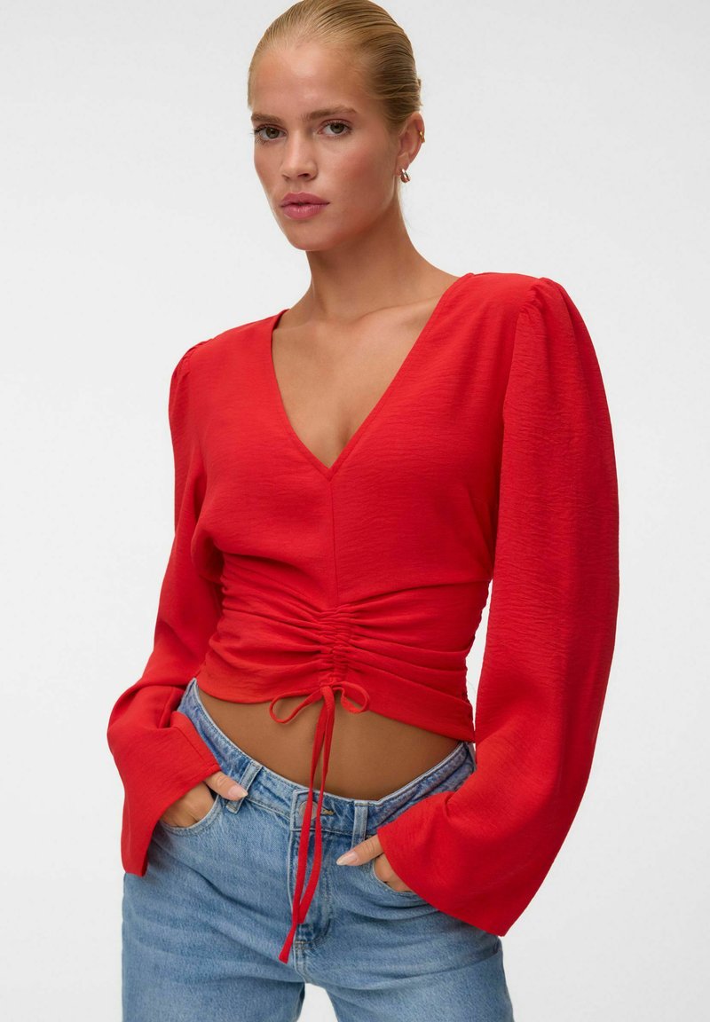 Femme aux cheveux blonds portant un haut rouge à col en V froncé à manches longues et un jean bleu, les mains dans les poches, sur un fond blanc uni.