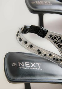 Schwarze High-Heel-Sandalen mit einem glänzenden silbernen Riemen, der mit schwarzen Strasssteinen verziert ist. Das Markenlogo "NEXT FOREVER COMFORT®" ist auf der Sohle sichtbar.