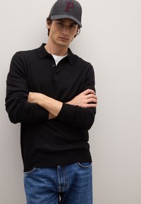 Schwarzes Polohemd mit langen Ärmeln und Kragen, kombiniert mit blauen Jeans. Ein graues Cap mit einem kastanienbraunen "P"-Logo sitzt auf dem Kopf des Models.