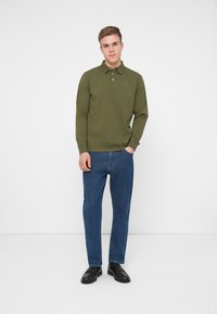 Oliwkowa, długiego rękawa koszulka polo z kołnierzykiem, w zestawieniu z prostymi niebieskimi dżinsami i czarnymi butami. Prosty, casualowy design.