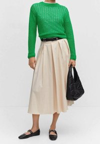 Grüner Strickpullover, cremefarbene Plisseerock, schwarzer Gürtel, schwarze gesteppte Handtasche und schwarze Ballerinas. Strukturiertes Material und geschichteter Design.