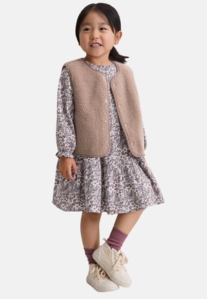 Beżowa kamizelka polarowa na sukience w kwiaty z długimi rękawami. Białe trampki i różowe skarpetki. Poza stojąca na białym tle.