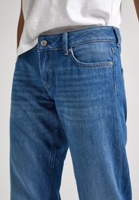 Jeans de mezclilla azul con corte recto, diseño de cinco bolsillos y herrajes de metal con una sutil textura desgastada. Se aprecian los pasadores para el cinturón y detalles de costura.