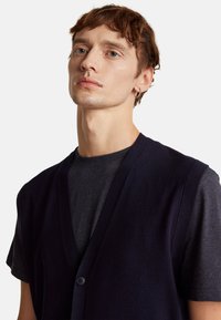 Chaleco de punto azul marino con un escote en V profundo, cierre de botones y sisas acanaladas, llevado sobre una camiseta de color carbón. Textura suave.