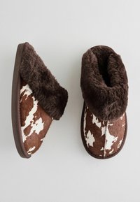 Faux-Fur-Hausschuhe mit einem braun-weißen Kuhfellmuster, einem weichen, strukturierten Kragen und einer dunkelbraunen Gummisohle für besseren Halt.