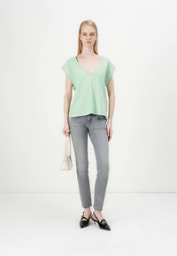 Top verde-menta de mangas curtas com decote em V, combinado com jeans skinny cinzentos. Modelo segura uma pequena mala branca e usa sapatilhas pretas com bico fino.