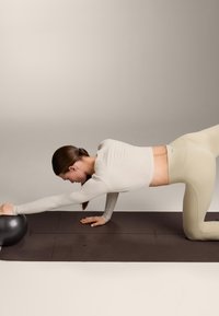 Žena v béžovém sportovním oblečení natahuje jednu ruku vpřed na jógové podložce a drží černý cvičební míč.