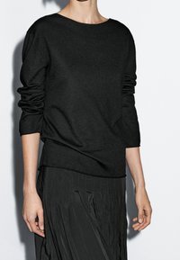 Musta pitkähihainen neulepaita, jossa on leveä pääntie ja pehmeä pinta, yhdistetty laskostettuun mustaan hameeseen. Asu on rento ja väljä.