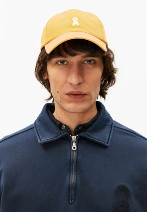 Giovane uomo con capelli castano medi, indossa un cappellino giallo e una giacca blu navy con zip, rivolto in avanti con un'espressione neutra.