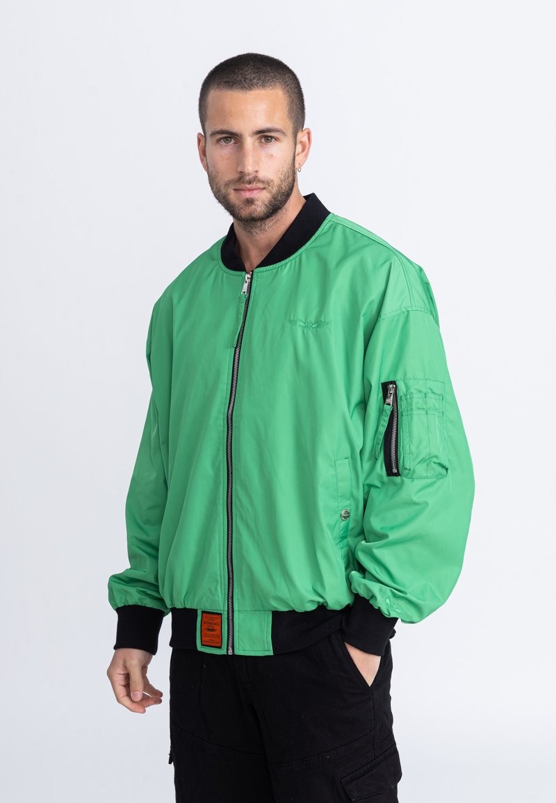 Veste bomber verte avec un col noir, fermeture éclair sur le devant et poche latérale. Dispose de poignets côtelés et d'un tissu texturé.