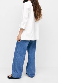 Weißes Langarmshirt mit hochgekrempelten Ärmeln und schwarzen Knöpfen, kombiniert mit locker sitzenden blauen Jeans aus Denim mit geradem Schnitt und verblasster Textur.
