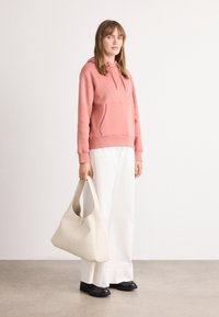 Felpa rosa con tasca frontale, abbinata a pantaloni bianchi a gamba larga. La modella tiene una borsa oversize color crema e indossa scarpe nere. Stile semplice e moderno.