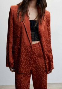 Veste et pantalon couleur rouille avec un motif texturé à pois. Le tissu semble lisse avec une légère brillance. Design à double boutonnage avec revers crantés.