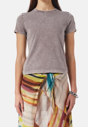 Femme portant un t-shirt gris délavé à manches courtes et un sarong coloré et à motifs noué à la taille, avec un bracelet chaîne au poignet droit.