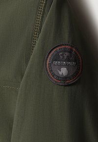 Olivgrön tyg med en texturerad yta. Har en rund svart lapp med orange detaljer och präglad text på vänster axel.