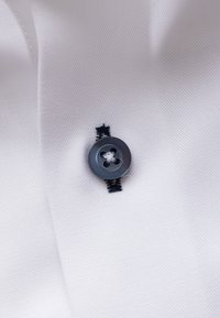 Bouton noir à quatre trous cousu avec du fil blanc sur un tissu blanc texturé avec une couture boutonnière noire.