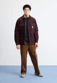 Brun kord fløjlsjakke med to brystlommer, parret med brune bukser og grå sneakers med blå striber. Enkel baggrund.