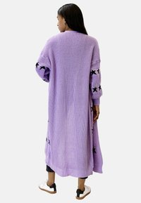 Larga chaqueta de punto lavanda con acentos de estrellas negras en las mangas, bordes acanalados y un diseño abierto. Llevada sobre pantalones negros y zapatos blancos.
